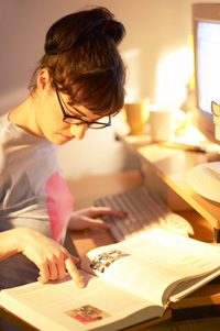 A female student studying from a textbook while sitting at a computer.